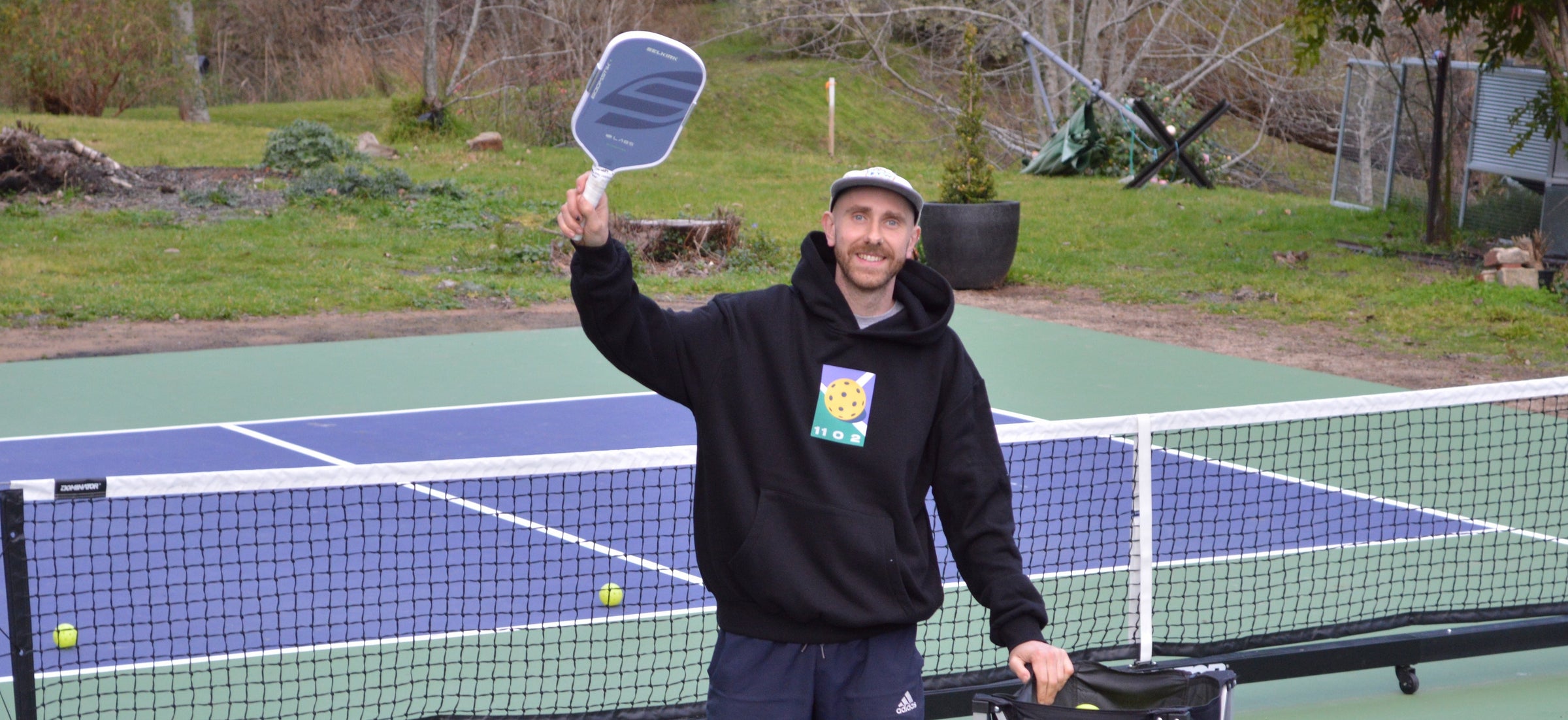 A man stands on a blue and green pickleball court holding a paddle and a wheeled black bag, with a net behind him and scattered yellow balls on the court.