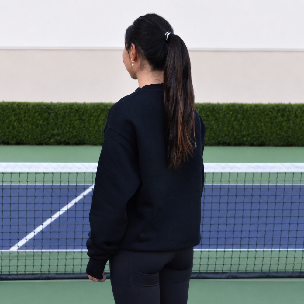 A woman stands with back facing, on a pickleball court wearing the black 11 0 2 dead dink crewneck.