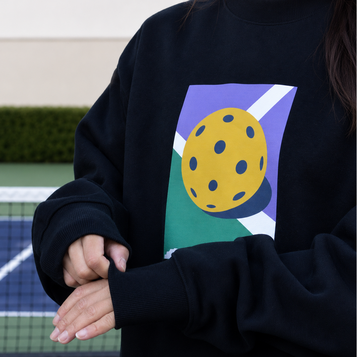 A girl wearing the oversized 11 0 2 dead dink black crewneck with a colourful graphic print of a yellow pickleball ball on a purple and green background, standing on a pickleball court.
