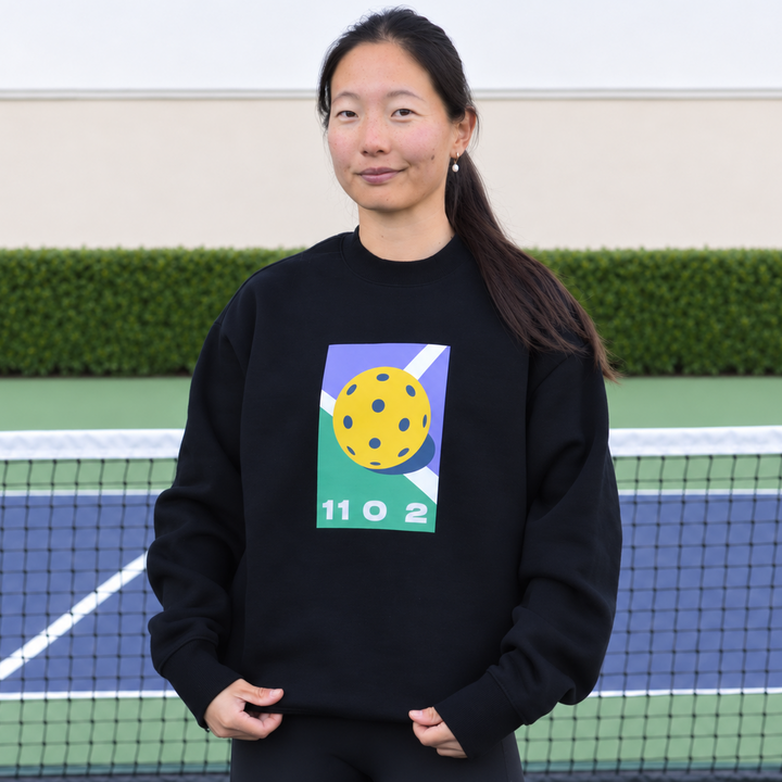 A woman stands front on, on a pickleball court wearing the black 11 0 2 dead dink crewneck.
