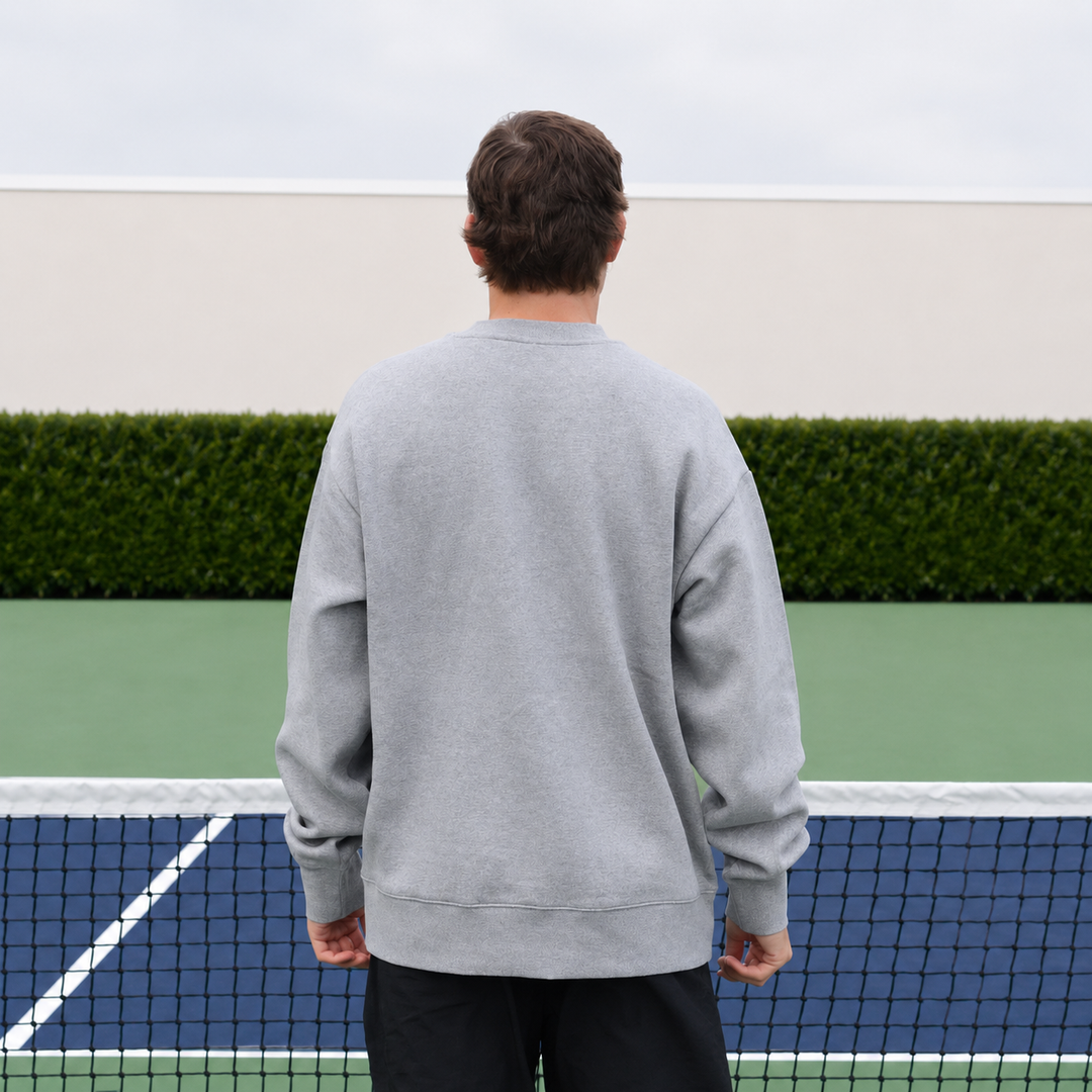 A man viewed from behind wearing a 11 0 2 dead dink crewneck in grey, standing on a pickleball court in front of a net and hedge.