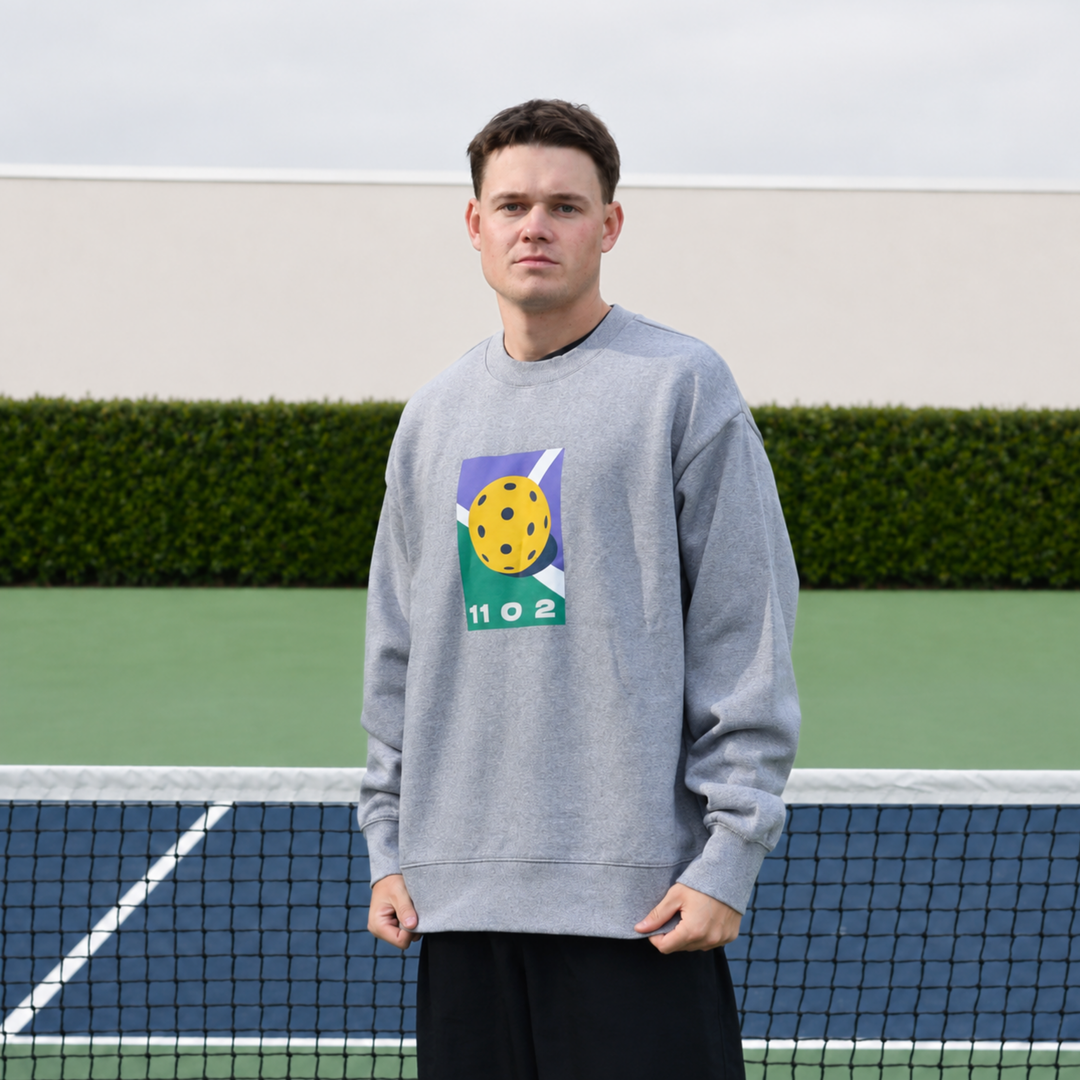 A man stands on a pickleball court wearing the 11 0 2 grey dead dink crewneck sweatshirt with a colourful graphic on the chest and black shorts.