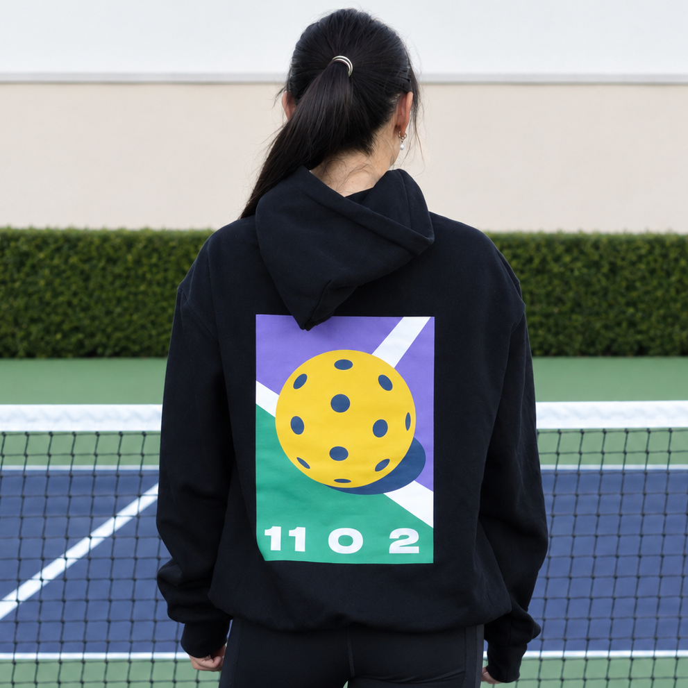 A woman viewed from behind wearing a black 11 0 2 dead dink hoodie. The print features a yellow pickleball on a purple, green, and white geometric background with the numbers &