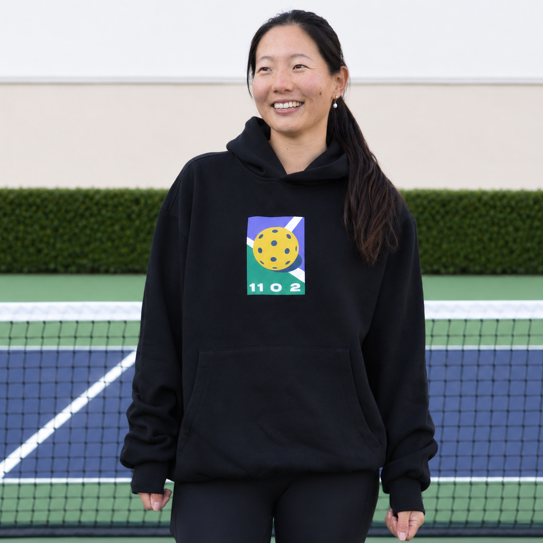 A woman stands on a pickleball court wearing a black dead dink hoodie from 11 0 2.