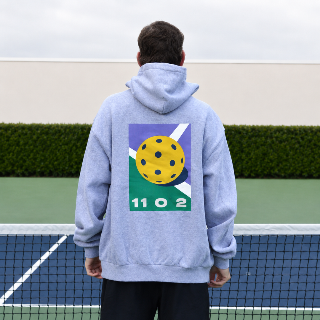 A man viewed from behind wearing a grey 11 0 2 dead dink hoodie with a large graphic print on the back, standing on a pickleball court with a net in the foreground and a hedge behind him.