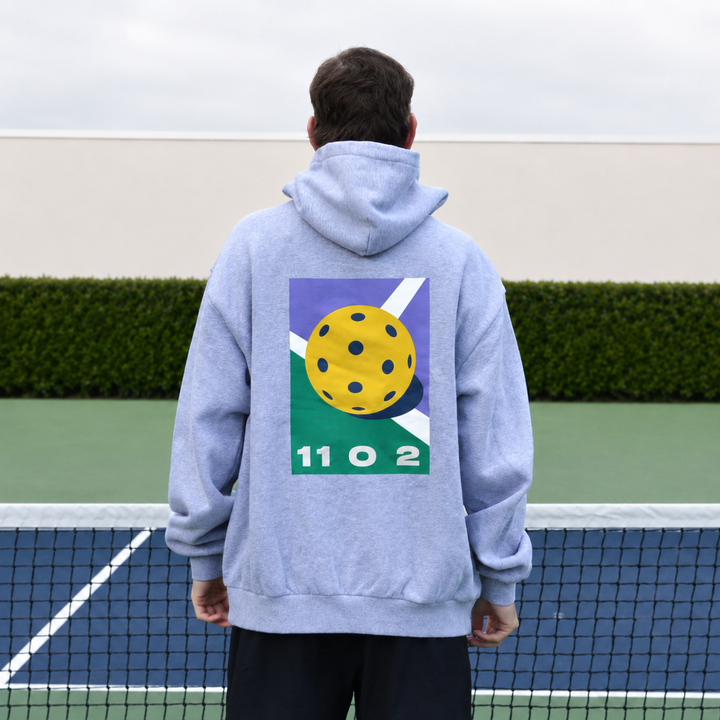 A man viewed from behind wearing a grey 11 0 2 dead dink hoodie with a large graphic print on the back, standing on a pickleball court with a net in the foreground and a hedge behind him.