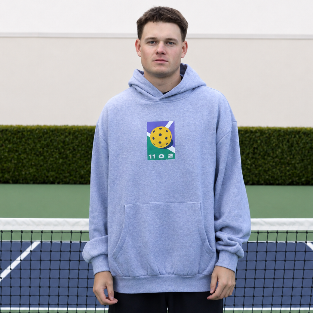 A man stands on a pickleball court wearing a grey 11 0 2 dead dink hoodie with a fun colourful print on the chest.