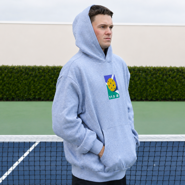 A man wearing an 11 0 2 grey dead dink hoodie stands on a pickleball court, with the hood over his head.
