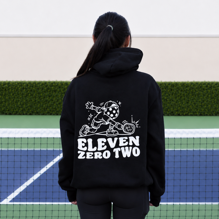 A woman standing on a green and blue pickleball court, viewed from behind, wearing a black 11 0 2 pickle head hoodie with a large white graphic and the text &