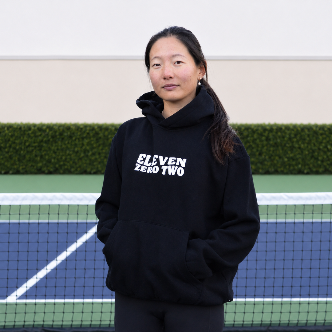 A woman stands on a blue and green pickleball court wearing a black 11 0 2 pickle head hoodie with &