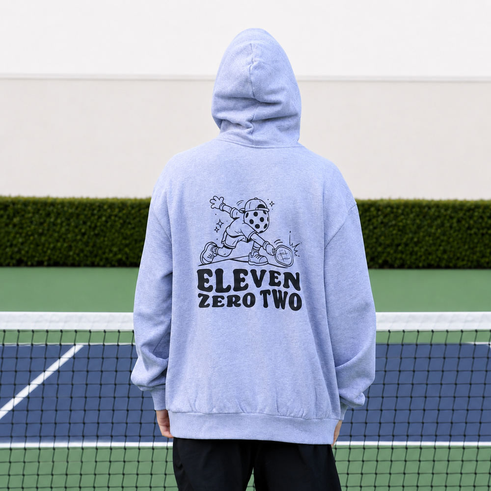 A man standing on a pickleball court, viewed from behind, wearing an oversized 11 0 2 pickle head hoodie in grey with a black graphic and the text 'ELEVEN ZERO TWO' printed on the back.