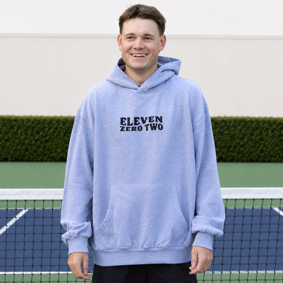 A smiling man wearing a grey 11 0 2 pickle head hoodie with 'ELEVEN ZERO TWO' printed in black on the chest, standing on a pickleball court in front of a net.