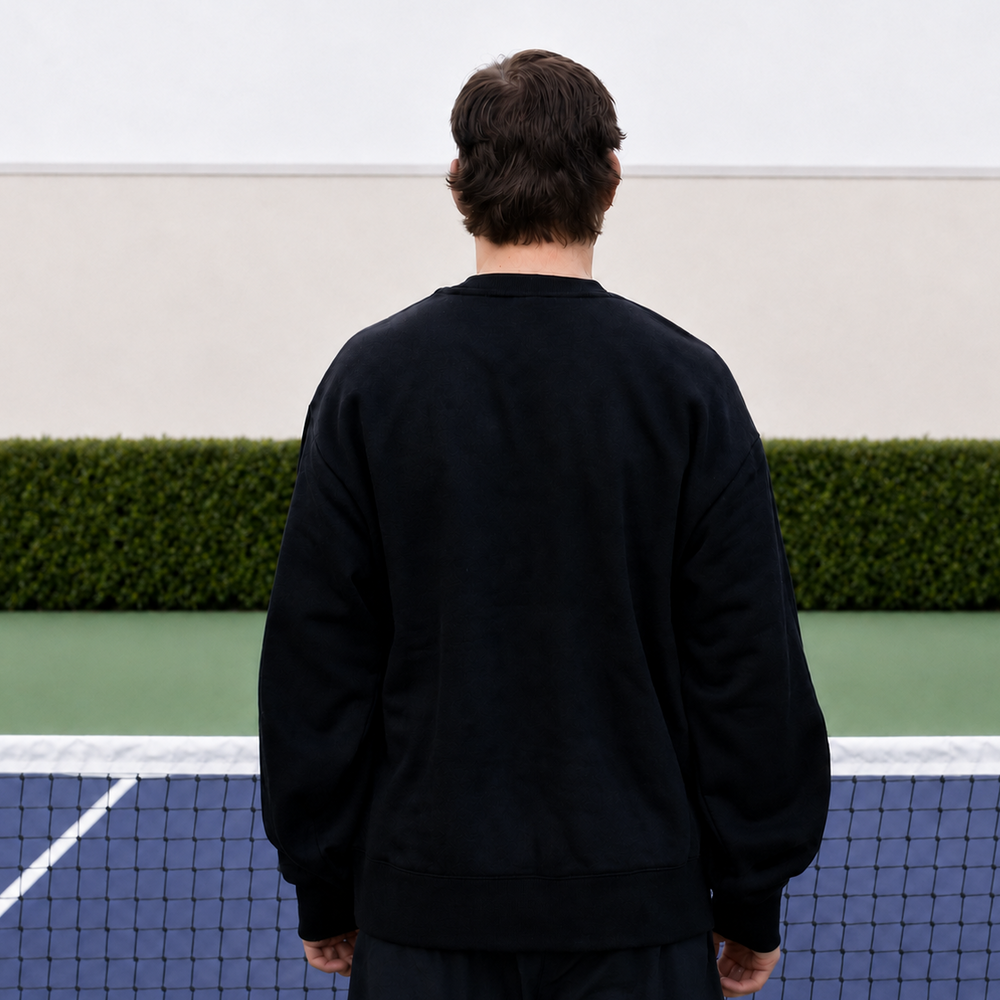 A man viewed from behind wearing a black 11 0 2 show court crewneck, standing on a blue and green pickleball court.
