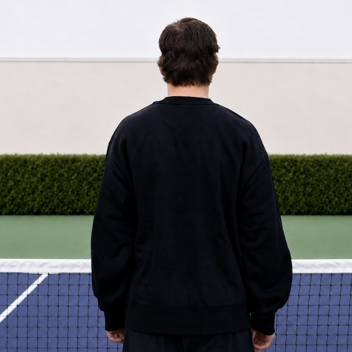 A man viewed from behind wearing a black 11 0 2 show court crewneck, standing on a blue and green pickleball court.