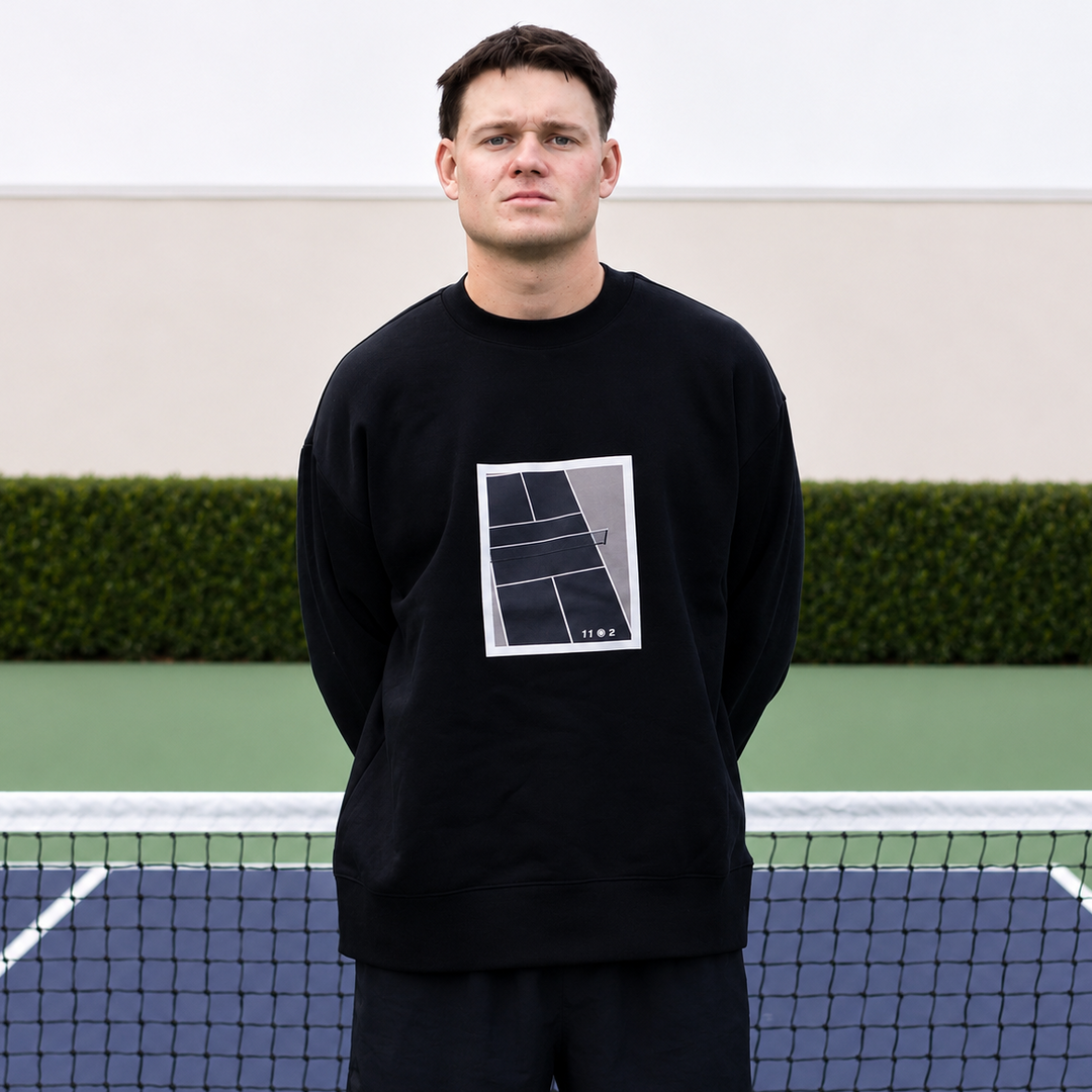 A man stands on a pickleball court wearing a black show court crewneck from 11 0 2.