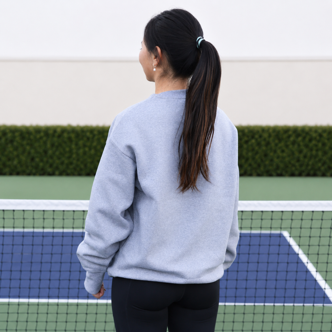 A woman viewed from behind wearing a grey 11 0 2 show court crewneck, standing on a blue and green pickleball court.