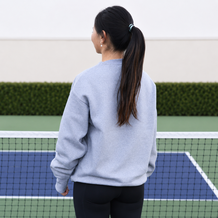 A woman viewed from behind wearing a grey 11 0 2 show court crewneck, standing on a blue and green pickleball court.