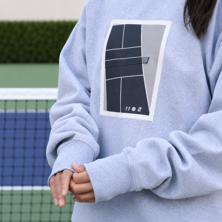 A woman wearing a light grey 11 0 2 show court crewneck jumper with a large pickleball court graphic on the chest, standing on a blue and green pickleball court.