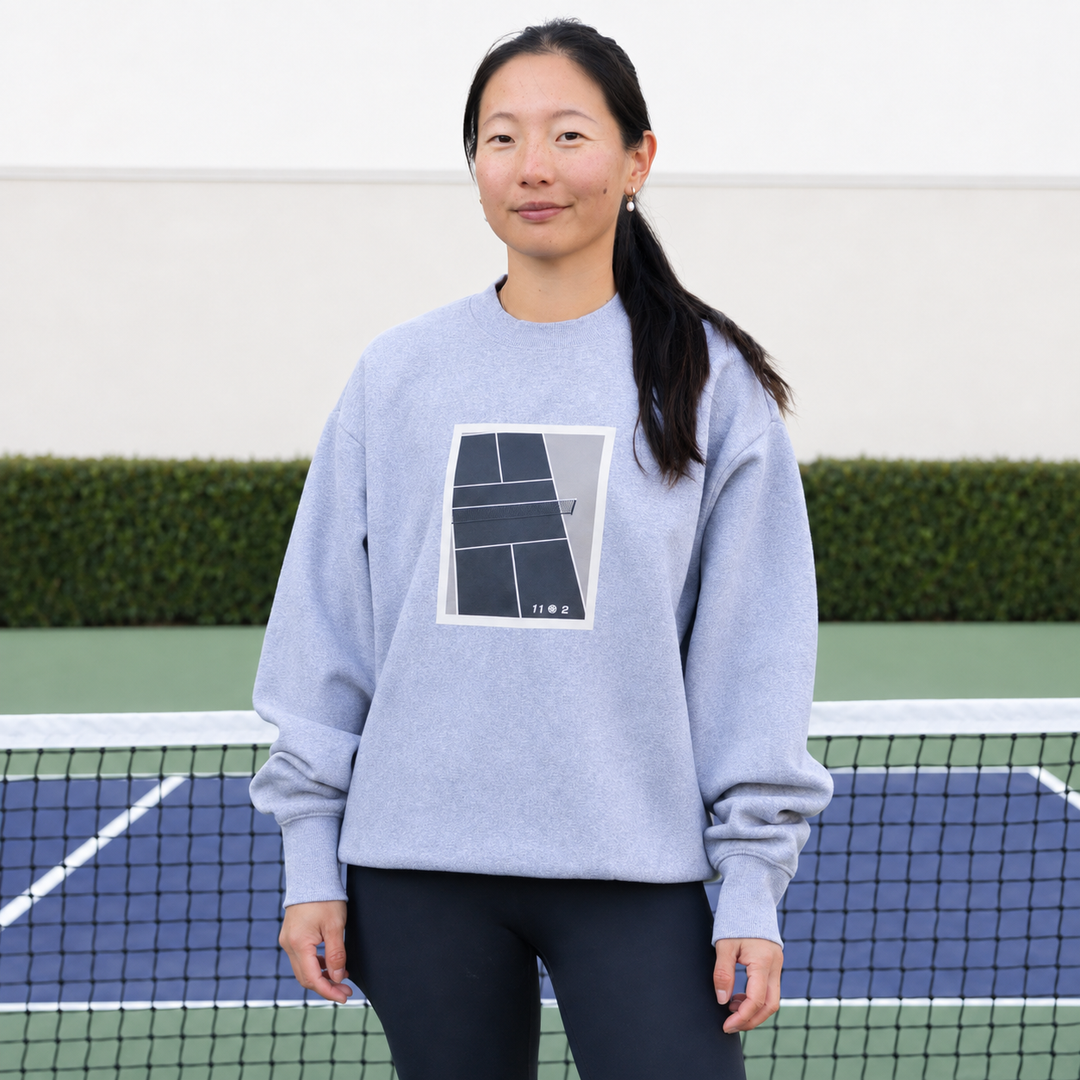 A woman stands on a blue and green pickleball court wearing a grey 11 0 2 show court crewneck.