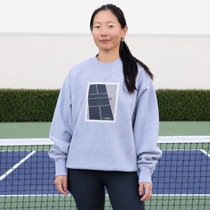 A woman stands on a blue and green pickleball court wearing a grey 11 0 2 show court crewneck.