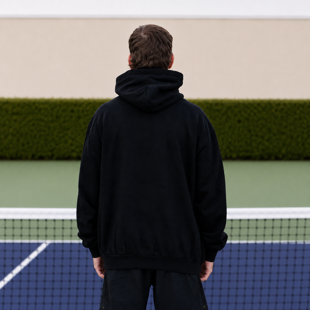 A man viewed from behind wearing an 11 0 2 show court hoodie in black, standing on a pickleball court with a net in front of him and a green hedge in the background.