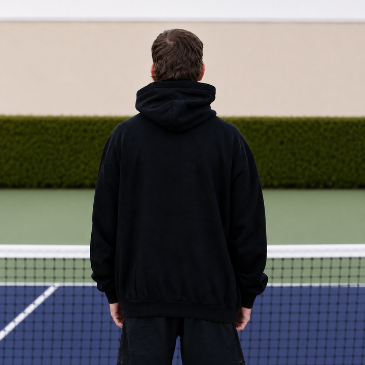 A man viewed from behind wearing an 11 0 2 show court hoodie in black, standing on a pickleball court with a net in front of him and a green hedge in the background.