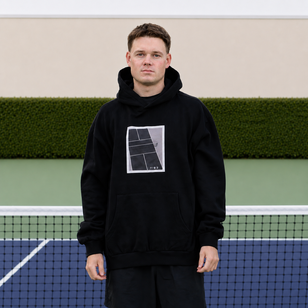 A man stands on a pickleball court wearing a black 11 0 2 show court hoodie.