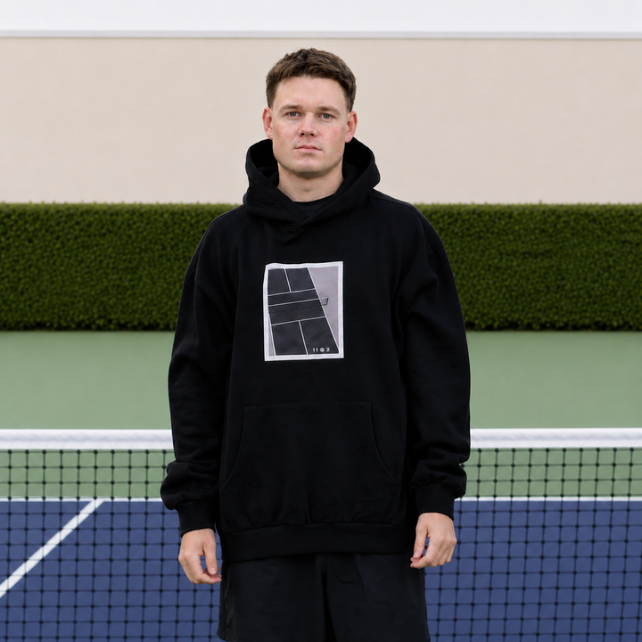 A man stands on a pickleball court wearing a black 11 0 2 show court hoodie.