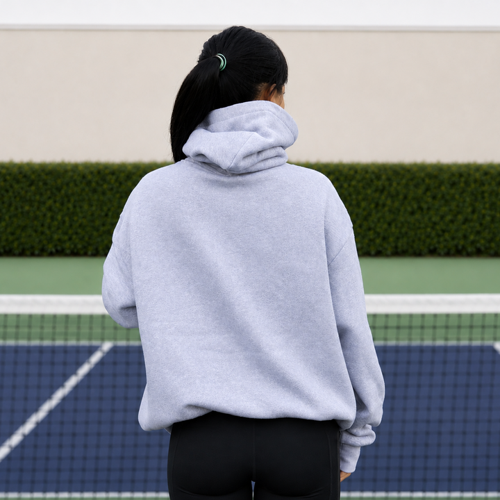 A woman viewed from behind wearing an oversized 11 0 2 grey show court hoodie standing on a pickleball court.
