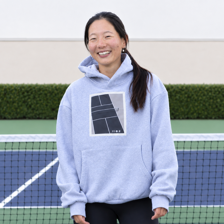 A woman stands on a pickleball court wearing an 11 0 2 show court hoodie in grey.
